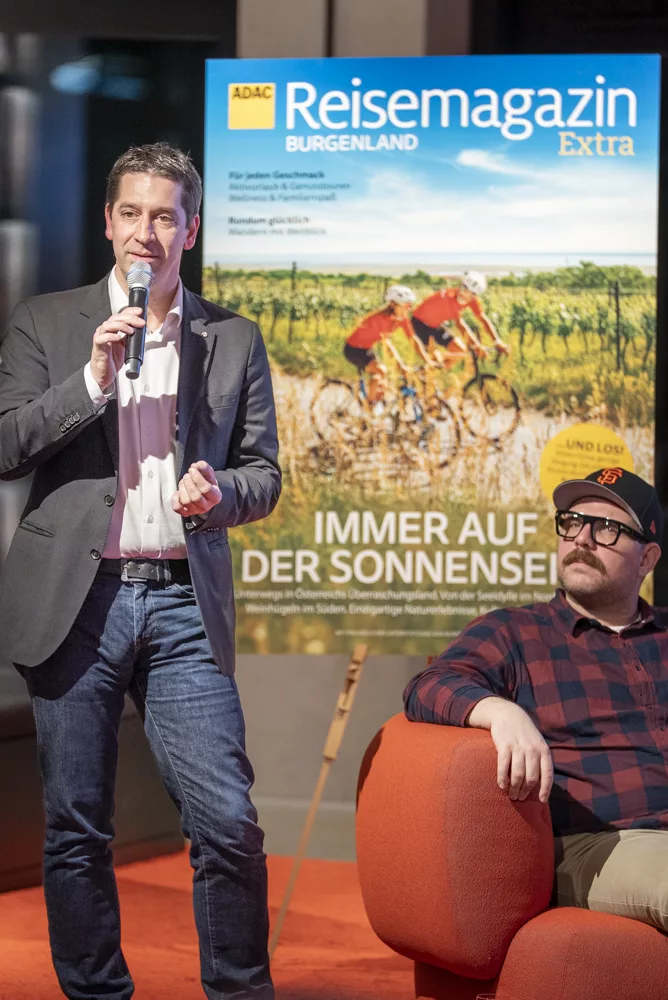 eventfotograf muenchen Mann mit Mikrofon spricht auf Veranstaltung, im Hintergrund Reisemagazin Burgenland
