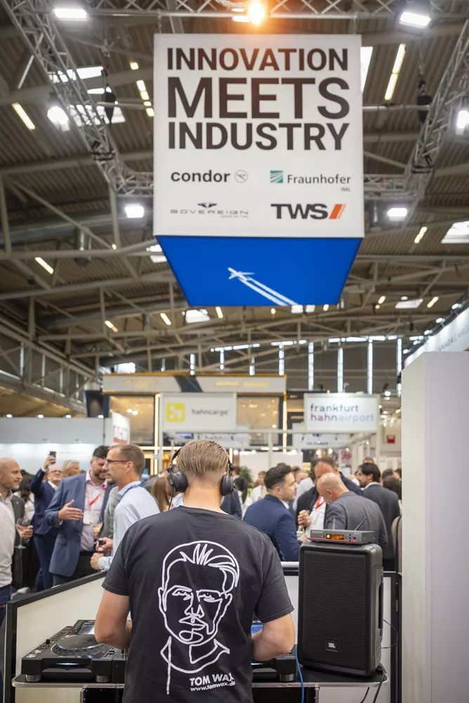 eventfotograf muenchen DJ spielt Musik auf Messe mit Menschen im Hintergrund und großem Banner